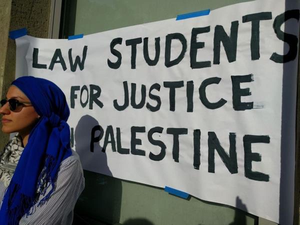 A student stands in front of a hand-painted sign reading "Law Students for Justice in Palestine."